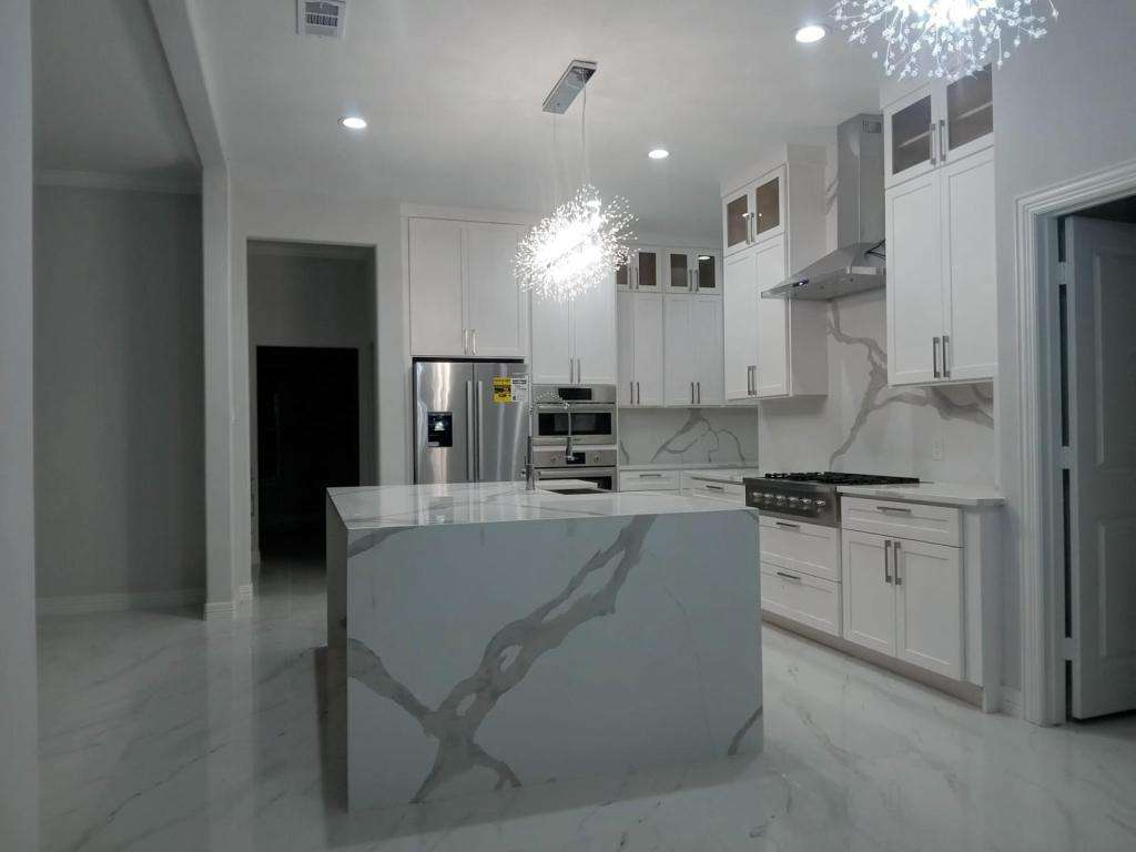 Modern kitchen with white cabinets, stainless steel appliances, marble-patterned island and countertops, built-in oven, and two contemporary pendant lights above the island.