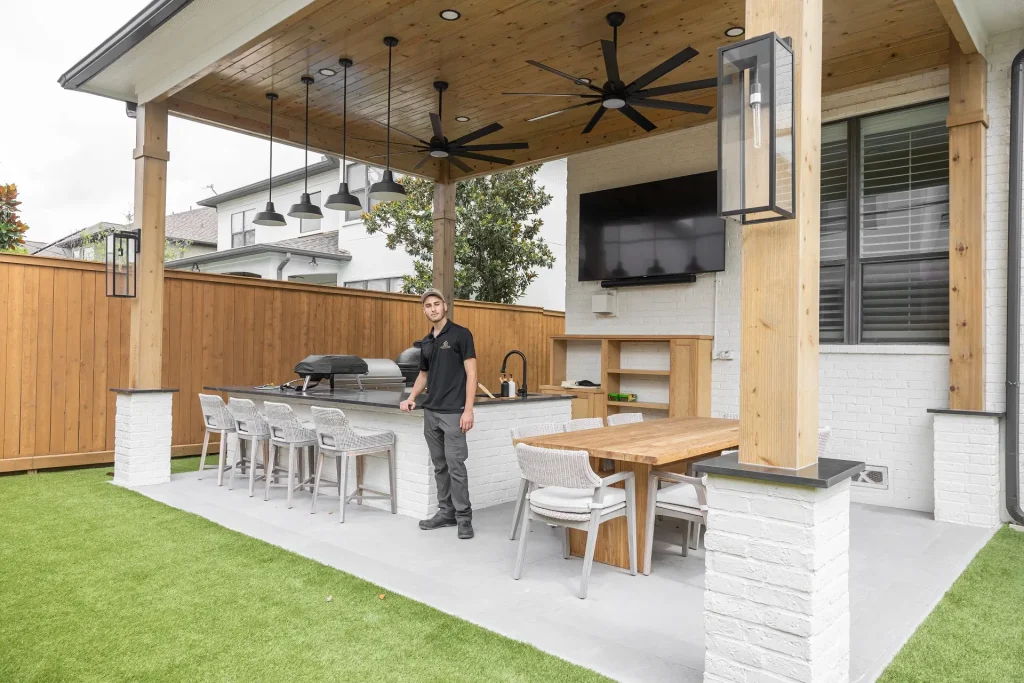 A man stands in a modern covered outdoor kitchen and dining area with a grill, bar seating, dining table, ceiling fans, TV, and wooden fence, on a patio with artificial grass.
